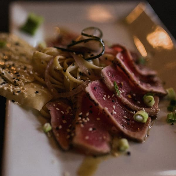 Seared tuna slices with sesame seeds, noodles, and green onions on a white plate.